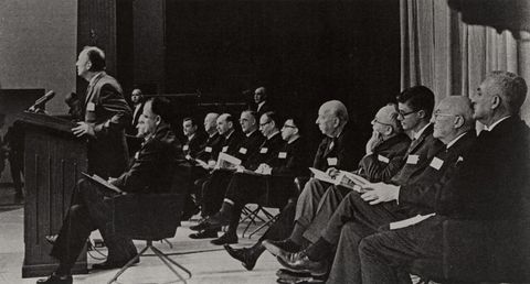 Speaker at a lectern with a row of seated men beside him at a 1964 press conference