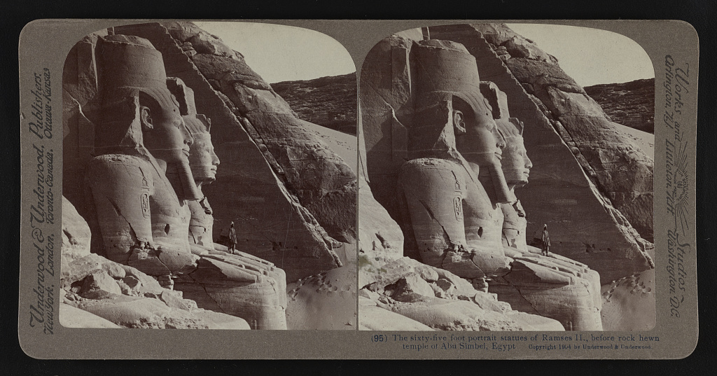 Stereograph of the colossal seated statues of Ramesses II at the entrance of the Great Temple at Abu Simbel, with a man standing at their feet for scale