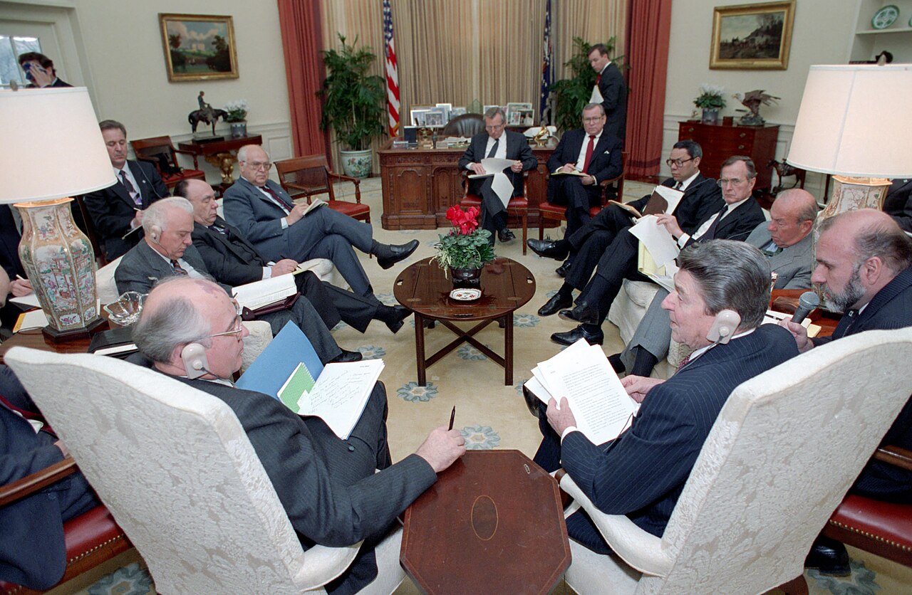 A group of U.S. and Soviet officials seated in armchairs in the Oval Office, several wearing translation earpieces; a desk and flags are behind them