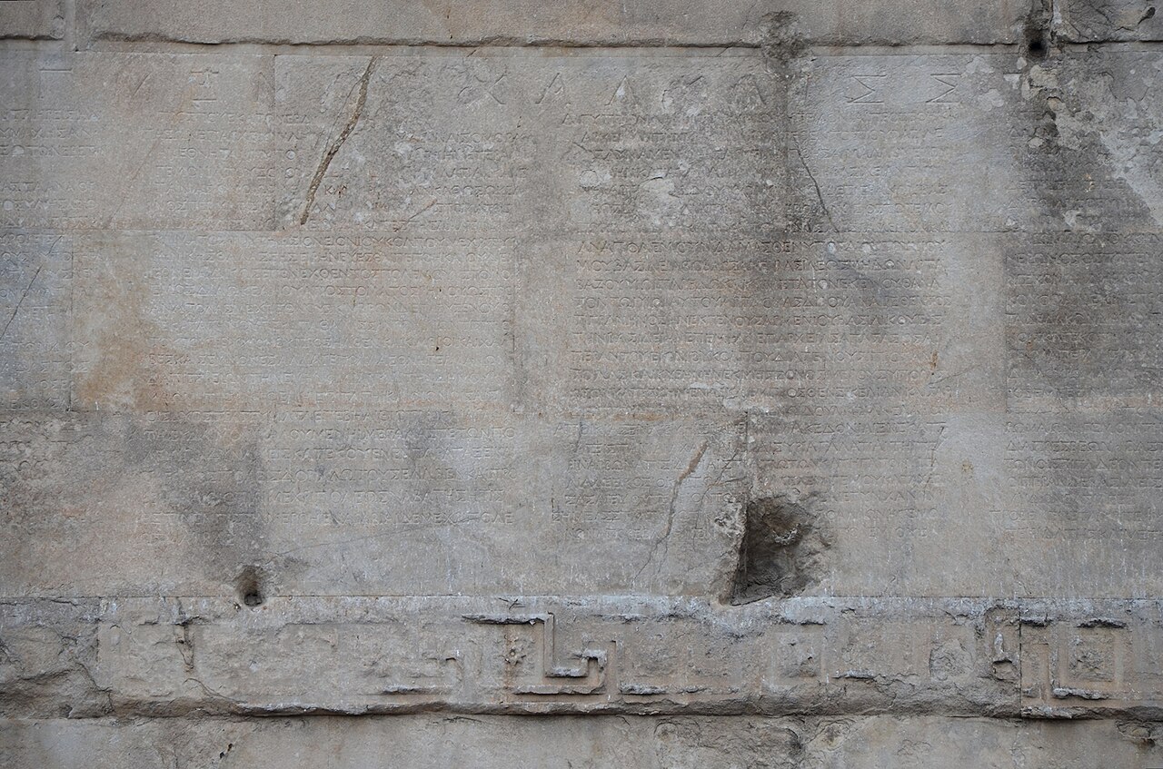 Photograph of a weathered marble wall covered in lines of Greek capitals above a carved meander band