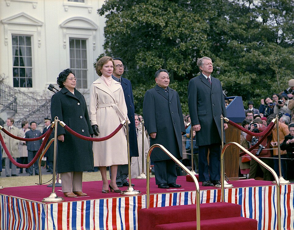 Deng Xiaoping and Jimmy Carter standing on a red-carpeted platform outside the White House beside Rosalynn Carter and Madame Zhuo Lin, with photographers behind a rope