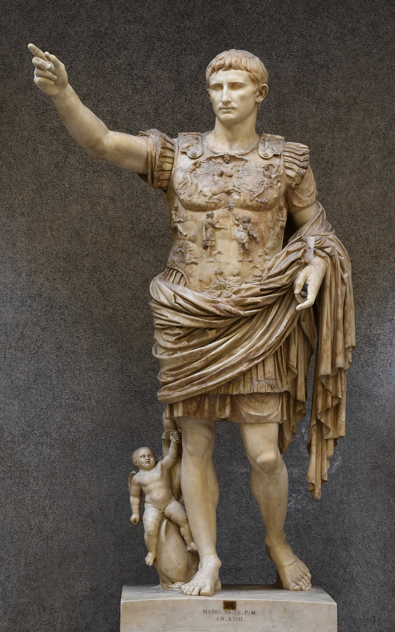 Marble statue of Augustus in a decorated breastplate, right arm raised, bare feet, with a small Cupid riding a dolphin at his right leg
