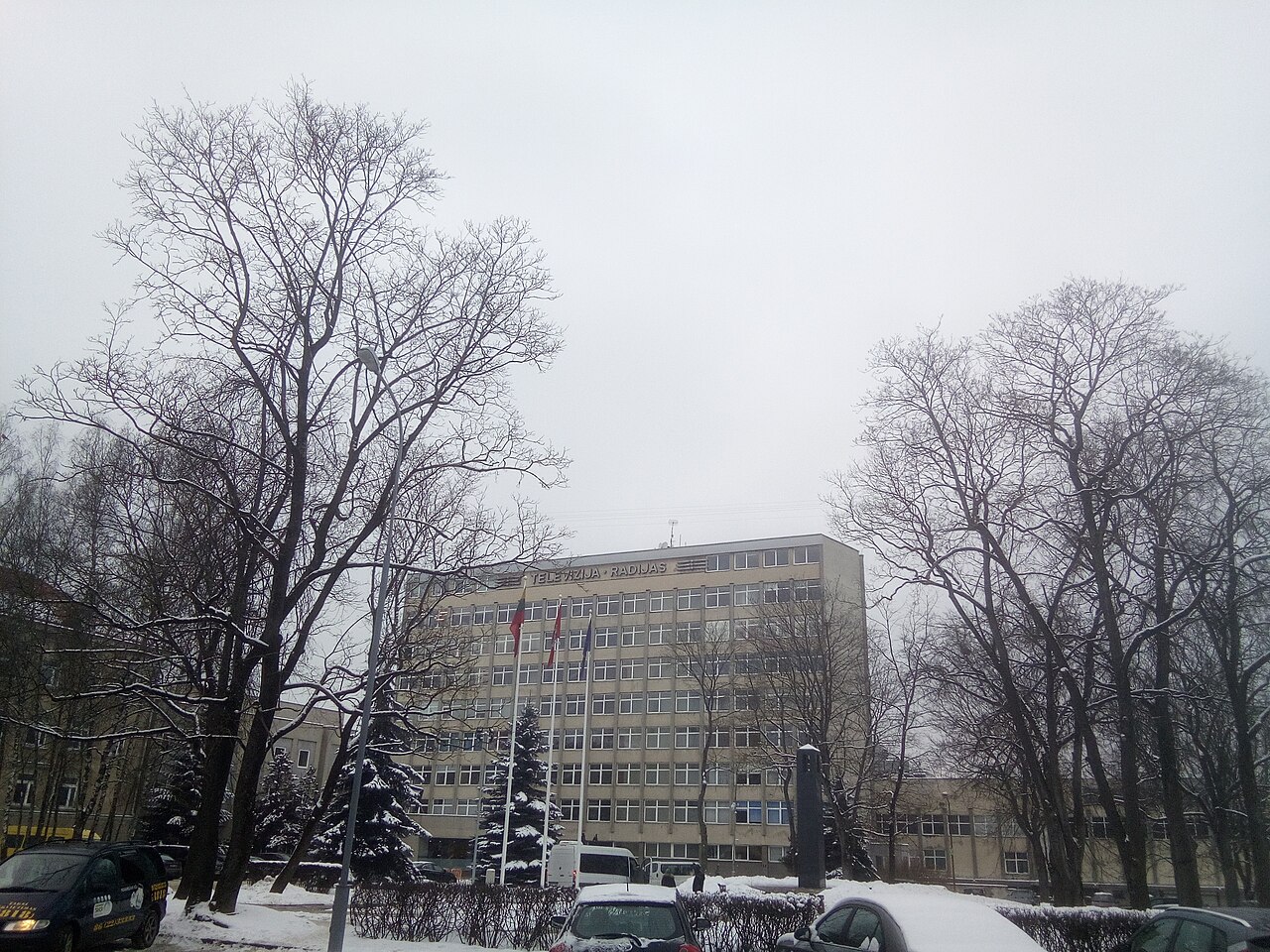 A multi-storey grey office building with the words Televizija and Radijas on its roof edge and three flagpoles in front, behind bare trees in snow
