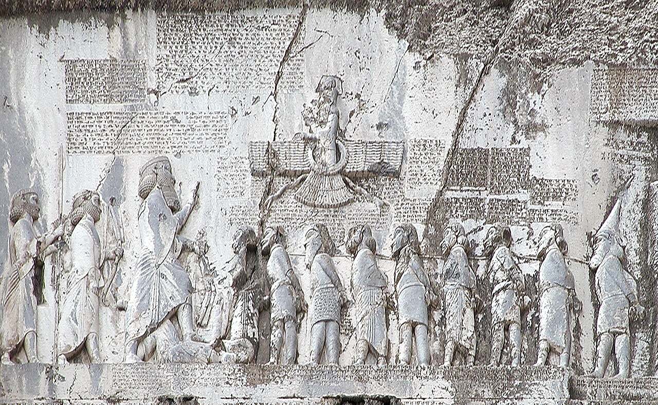 The Behistun relief: Darius, with two attendants behind him, stands with his foot on a prostrate figure, facing a line of bound captives roped at the neck; above them a winged figure in a disc