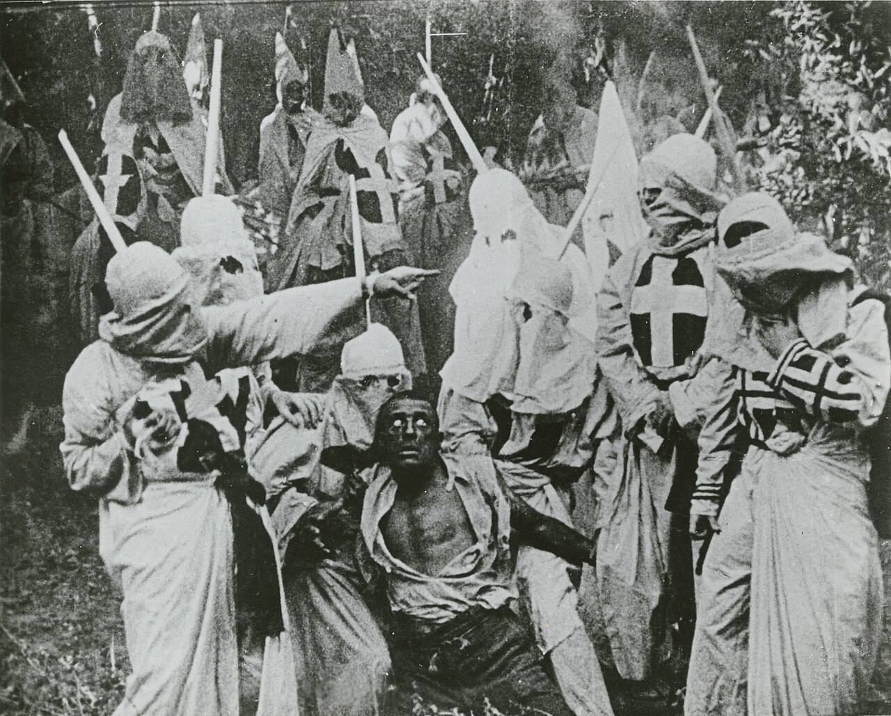 A publicity still from The Birth of a Nation: robed and hooded Klansmen with crosses on their chests stand around a seated man in torn shirt, a white actor in blackface playing the character Gus