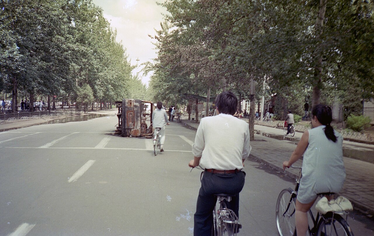A burned-out vehicle lies on its side on Zhongguancun street in Beijing's Haidian district as cyclists pass, June 1989