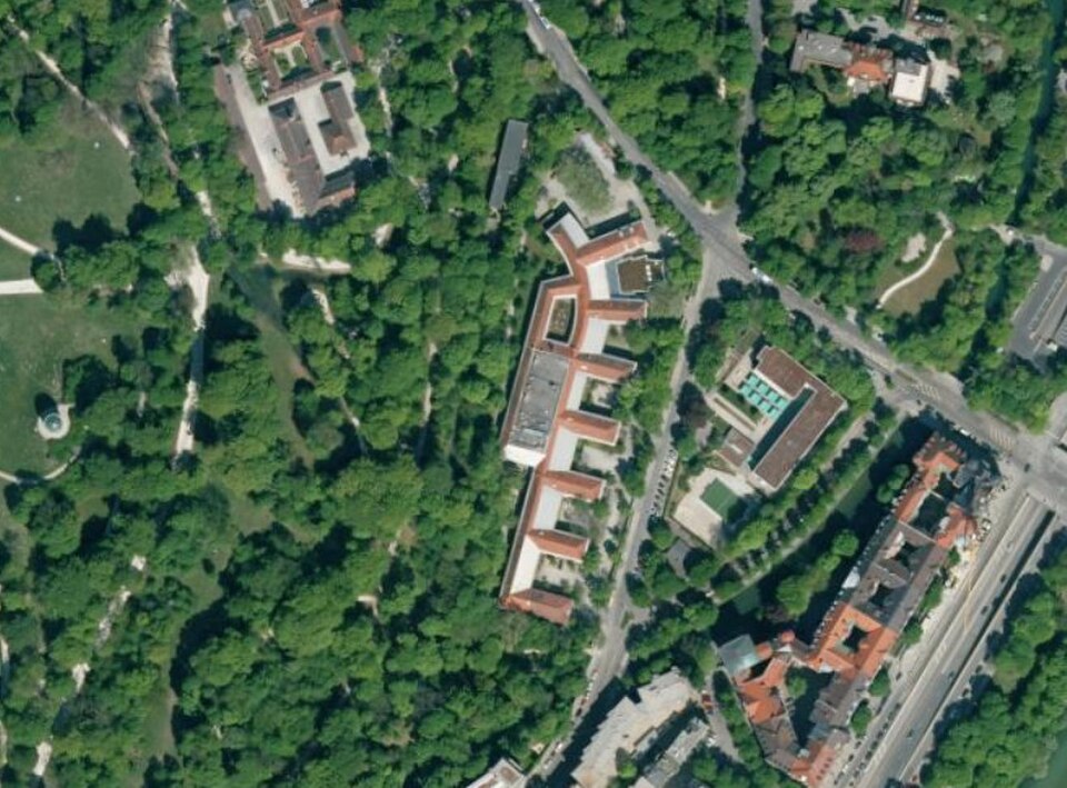 Aerial photograph of a long, angled office building with red-tiled roof beside the Englischer Garten in Munich