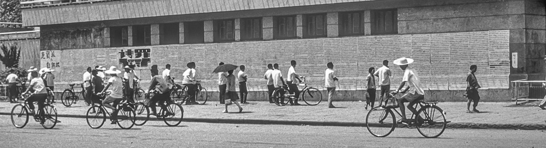A wall covered with handwritten big-character posters in Beijing, August 1976, with passers-by on bicycles