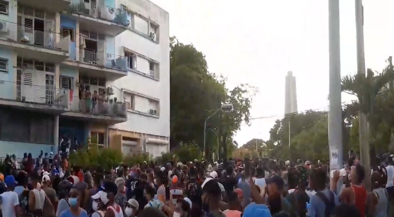 Crowd of people filling a street in Havana beside apartment blocks, with a tall monument in the background, in a still from a video