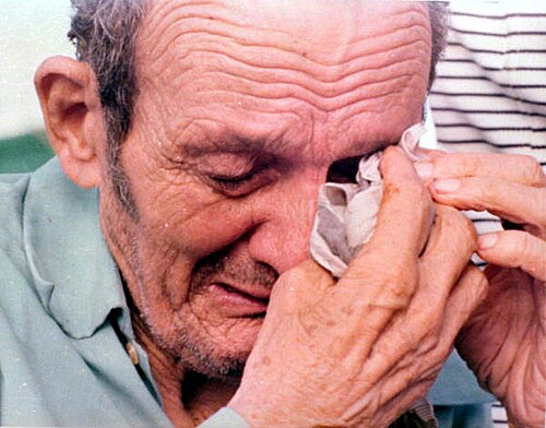 Close-up of an elderly man with his eyes closed, wiping one eye with a cloth held in his hand