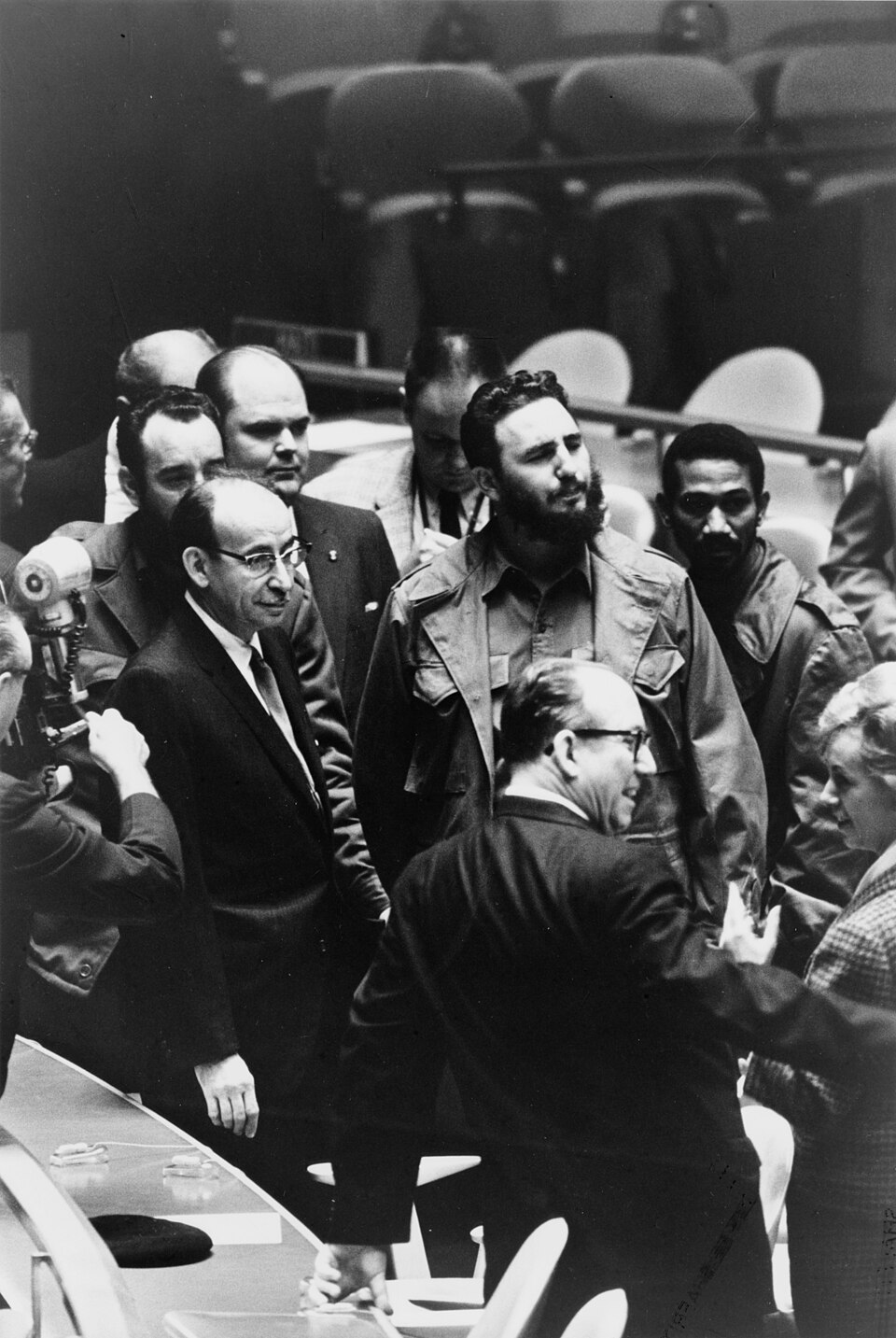 Fidel Castro in olive fatigues standing among men in suits on the floor of the United Nations General Assembly hall, a photographer at left