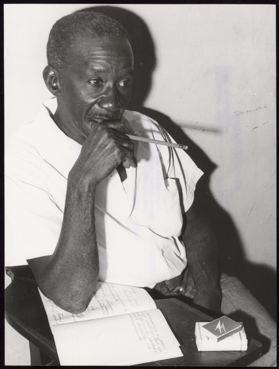 Black-and-white portrait of an older man in a white shirt at a school desk, a pencil at his lips, an open handwritten notebook and a cigarette pack in front of him
