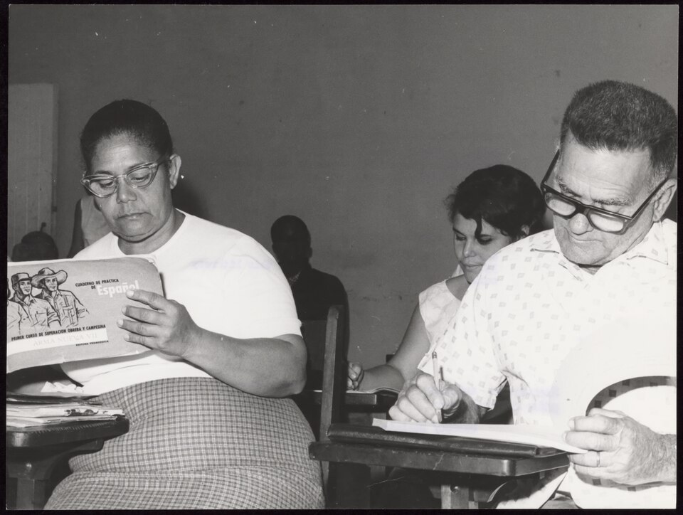 Two adults writing at school desks in a classroom, one holding a copybook marked Cuaderno de practica de Espanol, Primer curso de superacion obrera y campesina
