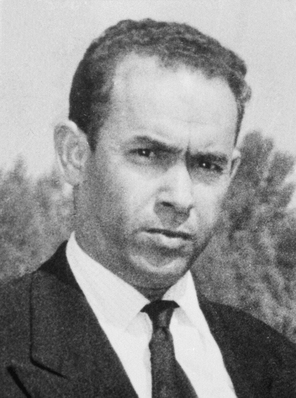 Black-and-white head-and-shoulders portrait of a man in a dark suit and tie with a stern expression