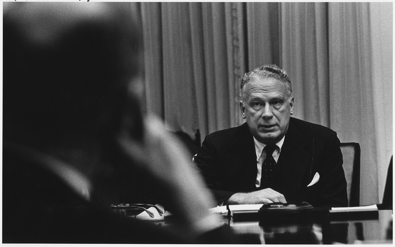 Black-and-white photograph of a man in a suit seated at a conference table speaking, with another person's head in the blurred foreground