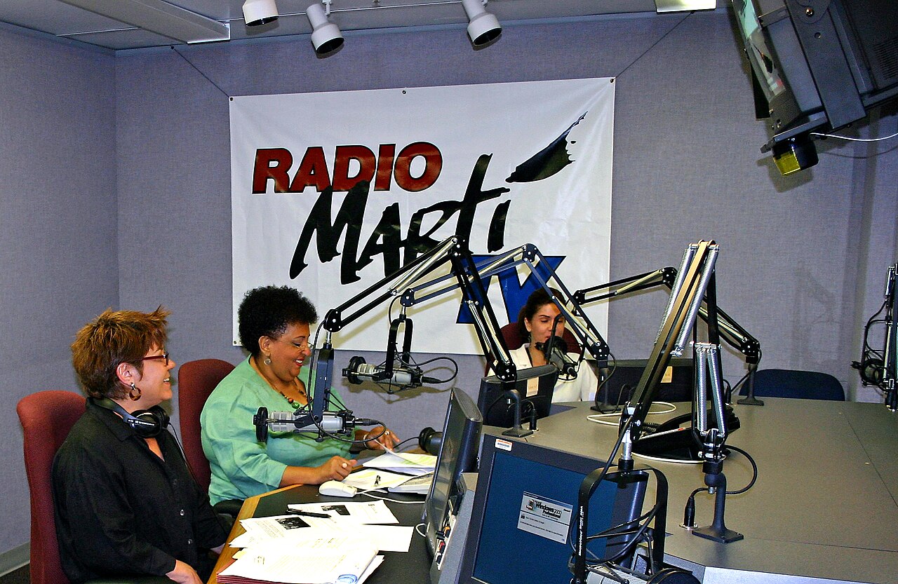 Two women at microphones and a third at a desk in a radio studio, with a large Radio Martí banner behind them