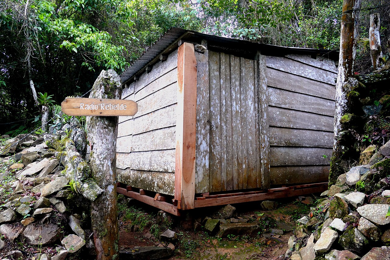 Small weathered wooden hut in forest at a trail junction, with a wooden sign reading Radio Rebelde