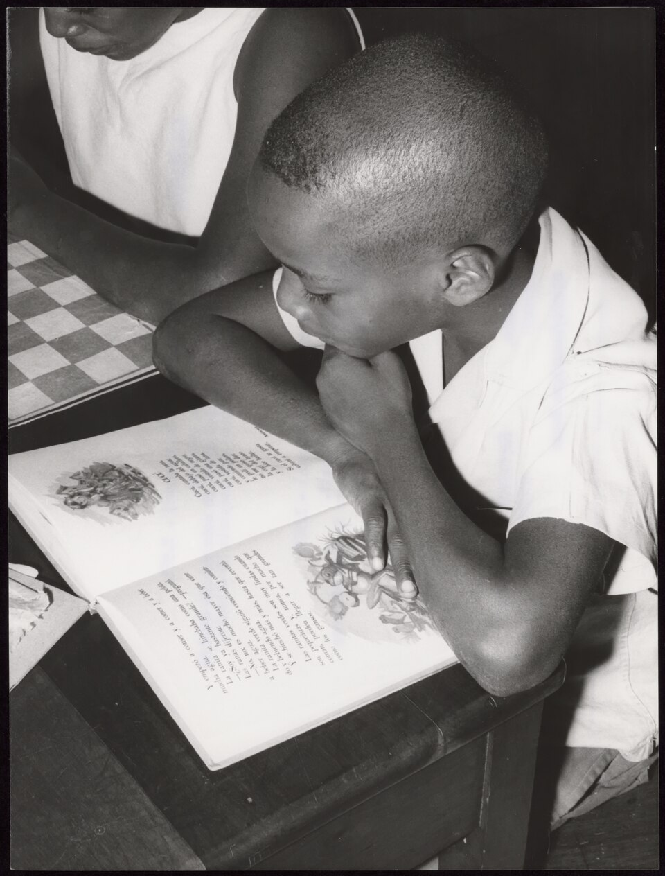 Black-and-white photograph of a boy in a white shirt leaning over an open illustrated book on a desk, reading