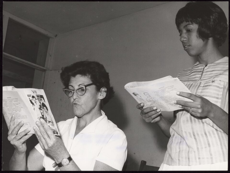 Two women in a classroom holding open exercise books, the older woman reading aloud with pursed lips