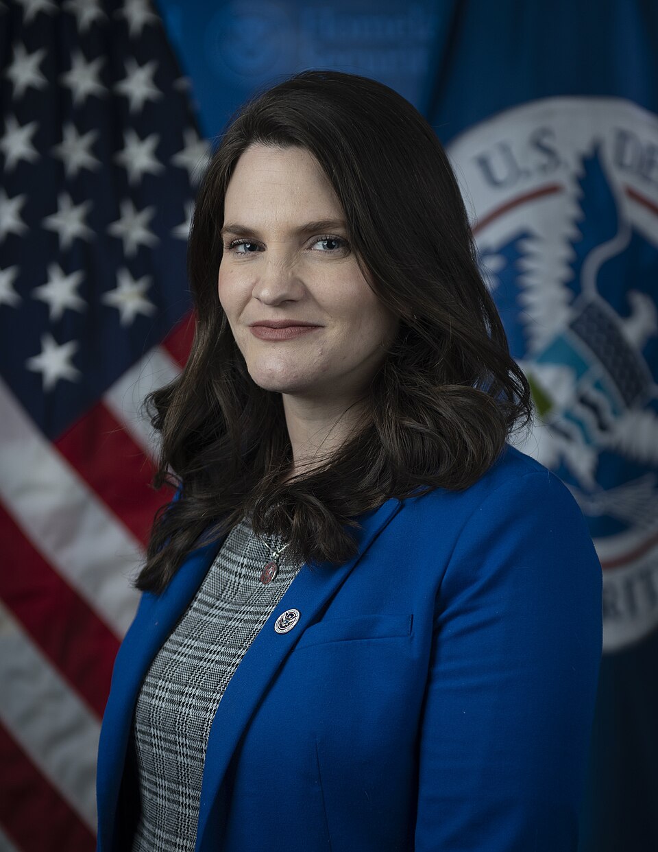 Head-and-shoulders portrait of a woman with dark hair in a blue jacket in front of a U.S. flag and the Department of Homeland Security seal