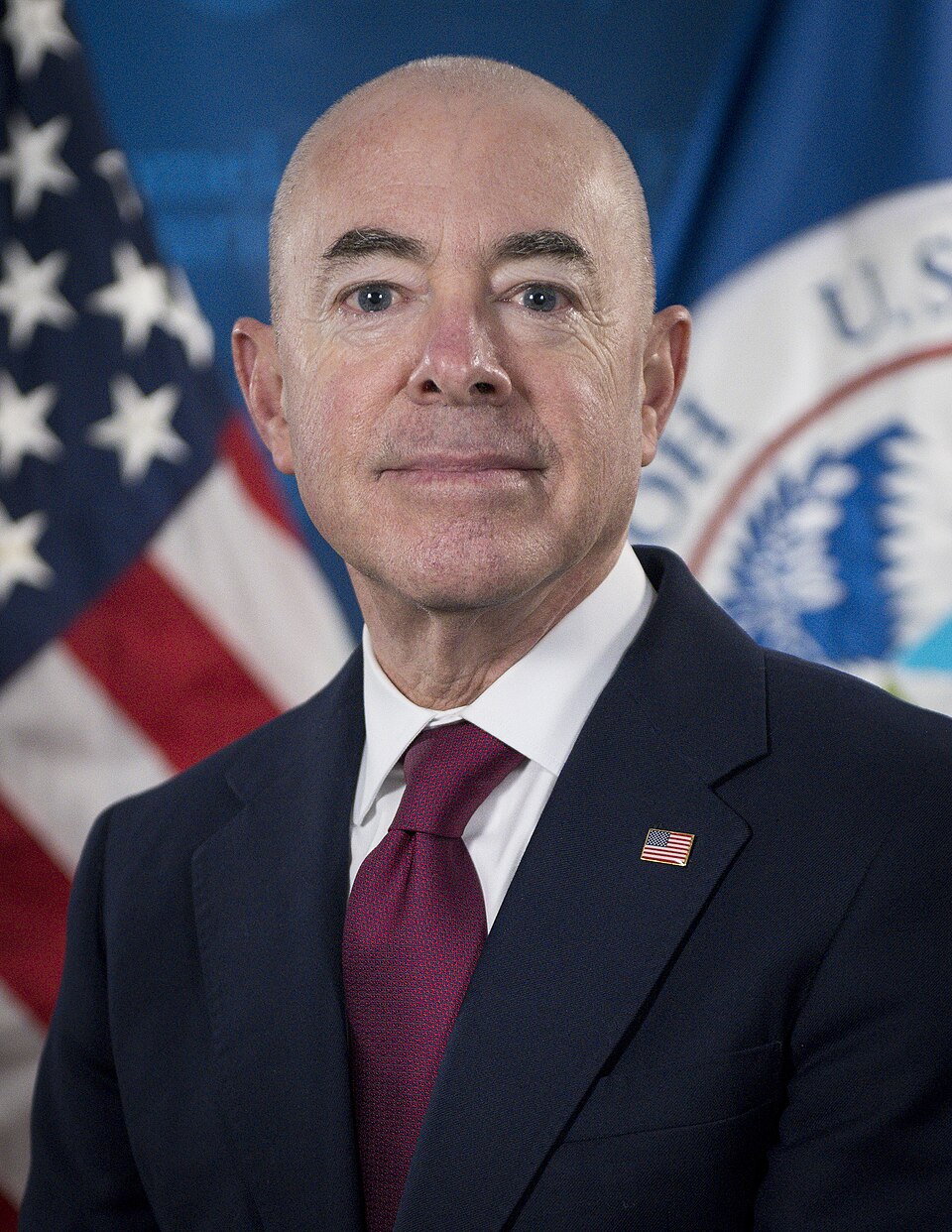 Head-and-shoulders portrait of a man in a dark suit and dark red tie in front of a U.S. flag and the Department of Homeland Security seal