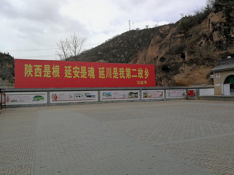 Large red banner with yellow Chinese characters above a paved plaza with wall panels, in front of a loess hillside at Liangjiahe village