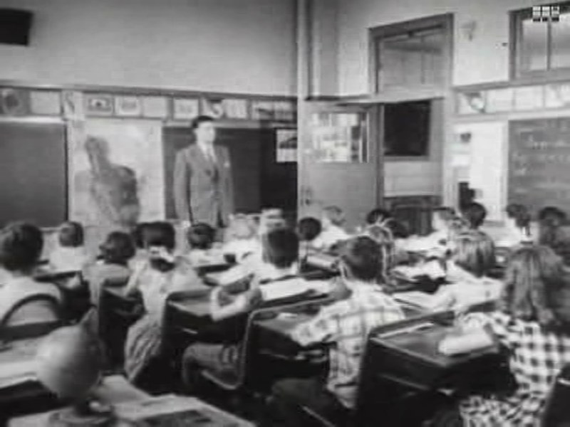 Frame from Duck and Cover: a teacher stands before rows of children at their desks in a classroom