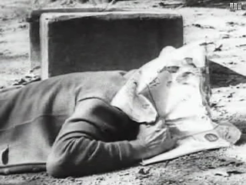 Frame from Duck and Cover: a boy lying face down on the ground behind a box, with a picnic blanket and newspaper over his head