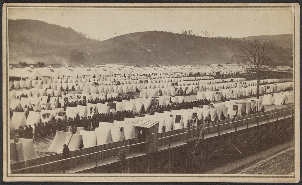 Photograph: rows of white tents and barracks inside a board fence at Elmira, New York, with lines of men between the tents and a guard walkway along the fence