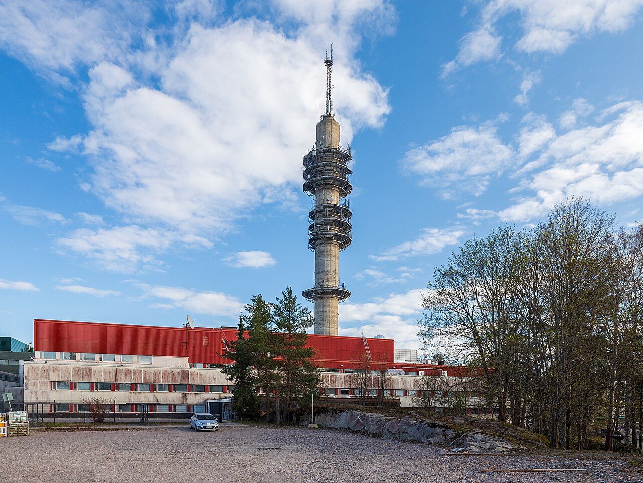 A low red and white studio building with a tall concrete transmission tower rising behind it under a blue sky