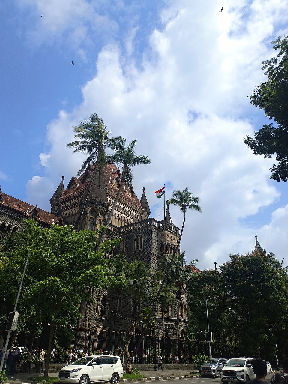 A dark stone Gothic Revival court building with turrets and a red-tiled roof, an Indian flag flying above palm trees and street traffic