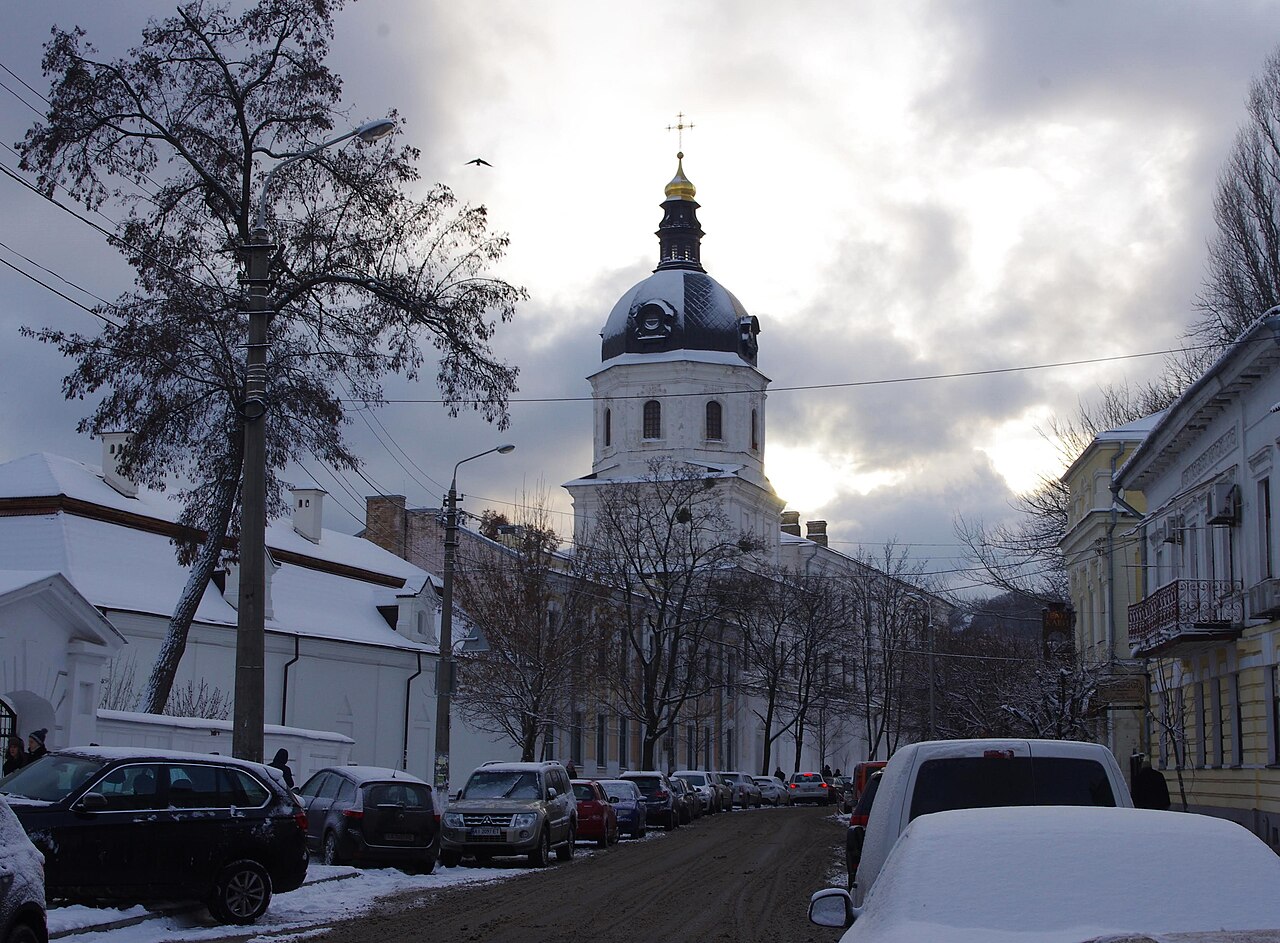 A snowy Kyiv street with a white bell tower topped by a black dome and gold cupola rising behind older buildings