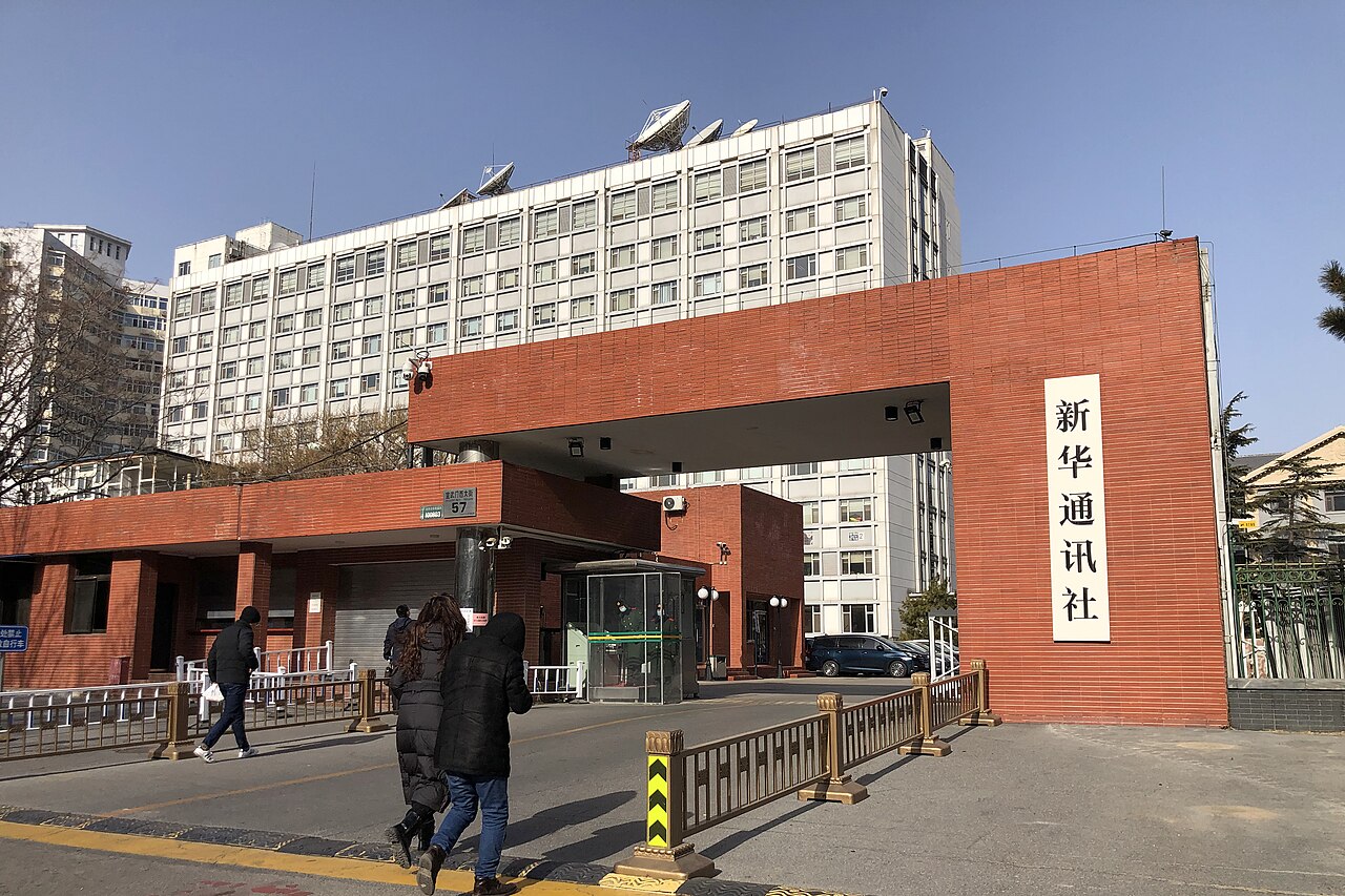 A brick gateway with a vertical sign reading 新华通讯社 (Xinhua News Agency) in front of a white multi-storey office building with satellite dishes on its roof
