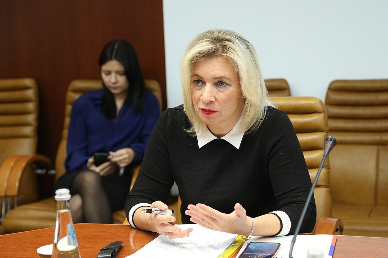 A woman with blonde hair seated at a wooden table with papers and a microphone, speaking, in a conference room