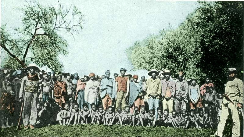 Hand-coloured postcard titled Kriegsgefangene Hottentotten: a group of men, women and children with two armed guards