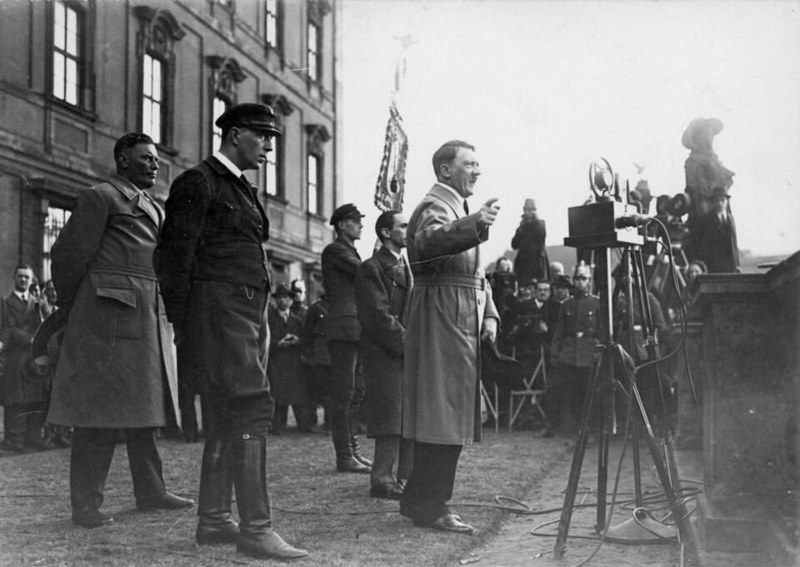 Photograph of Adolf Hitler in a trench coat speaking at a microphone on an open-air platform, with uniformed men, press photographers and a film camera around him