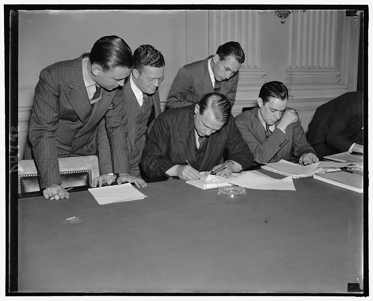 Five men in suits around a committee-room table; one seated man signs papers while others look on