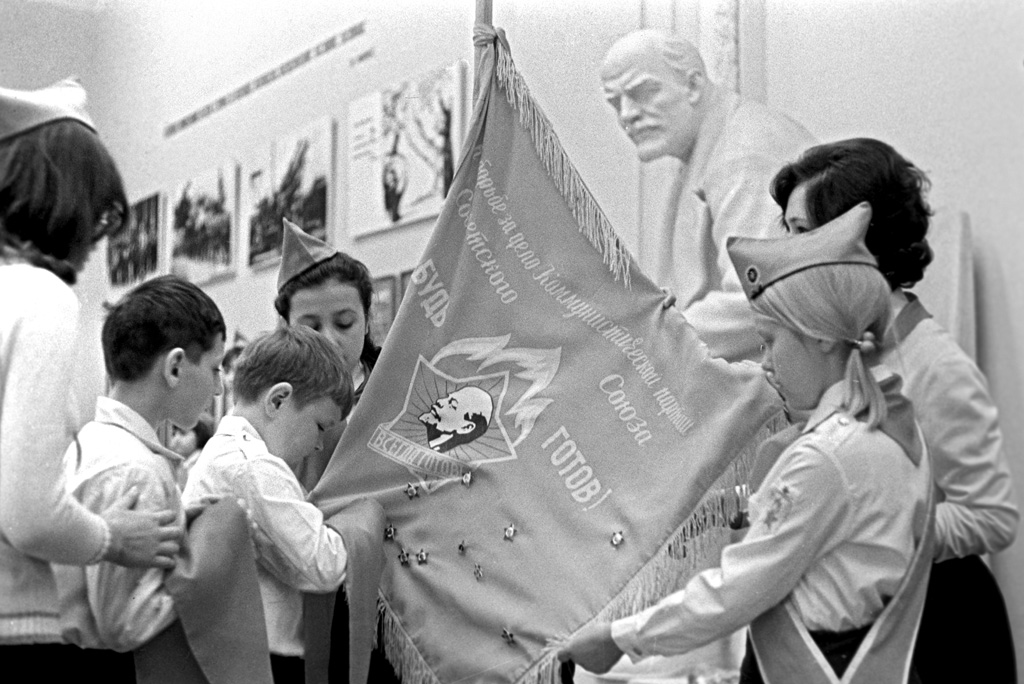 Photograph of a black-and-white 1981 press image: two boys without Pioneer neckerchiefs stand beside older girls in Young Pioneer uniform holding a red banner bearing a profile of young Lenin and the slogan "Be ready! For the cause of the Communist Party of the Soviet Union, be ready!", in front of a bust of Lenin at the Central Lenin Museum, Moscow