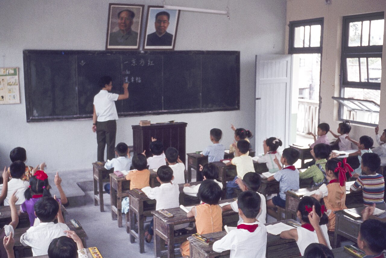 Primary-school classroom in China in 1978: a teacher writing at the blackboard beneath two framed portraits, pupils in red neckerchiefs raising their hands