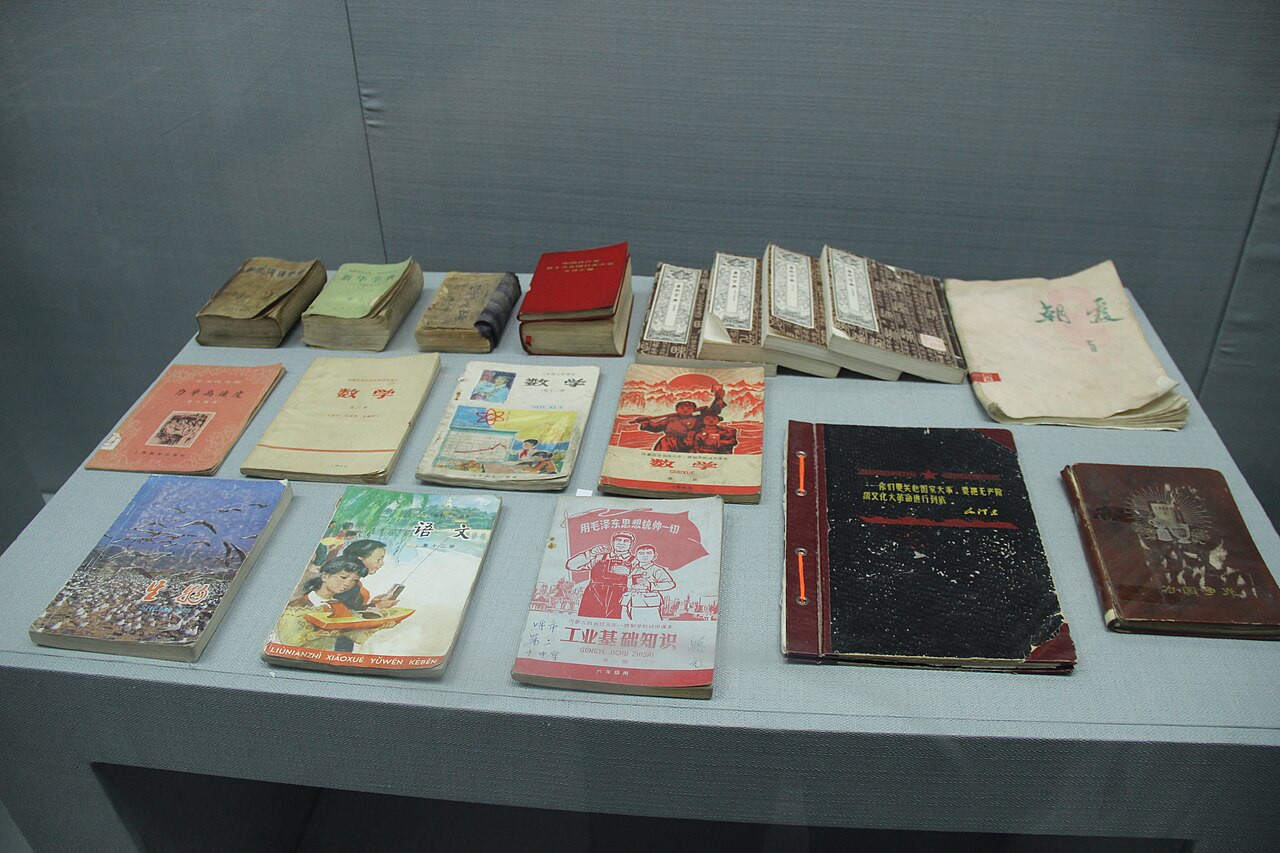 Museum display case of Cultural Revolution-era textbooks and notebooks, including a reader whose cover bears a slogan about Mao Zedong Thought