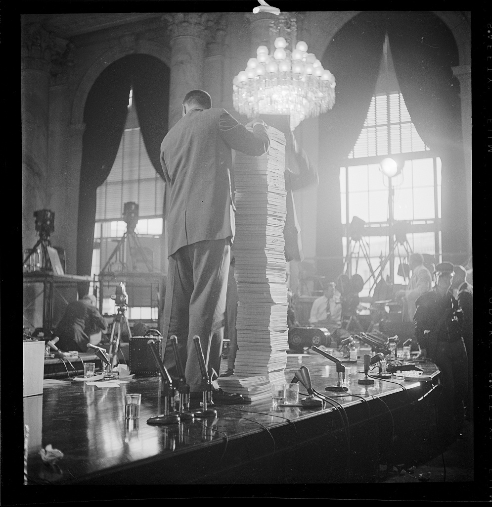 A man stands on the committee table beside a tall stack of documents during the Army-McCarthy hearings, 1954