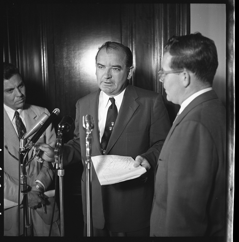 Senator Joseph McCarthy holding a sheet of papers and speaking before several microphones, with two men beside him