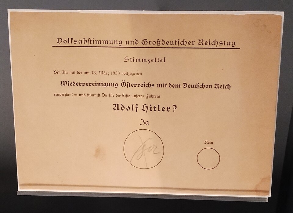 Photograph of a ballot paper reading "Volksabstimmung und Großdeutscher Reichstag": "Are you in agreement with the reunification of Austria with the German Reich carried out on 13 March 1938 and do you vote for the list of our Führer Adolf Hitler?" with a large circle marked "Ja", crossed, and a much smaller circle marked "Nein"