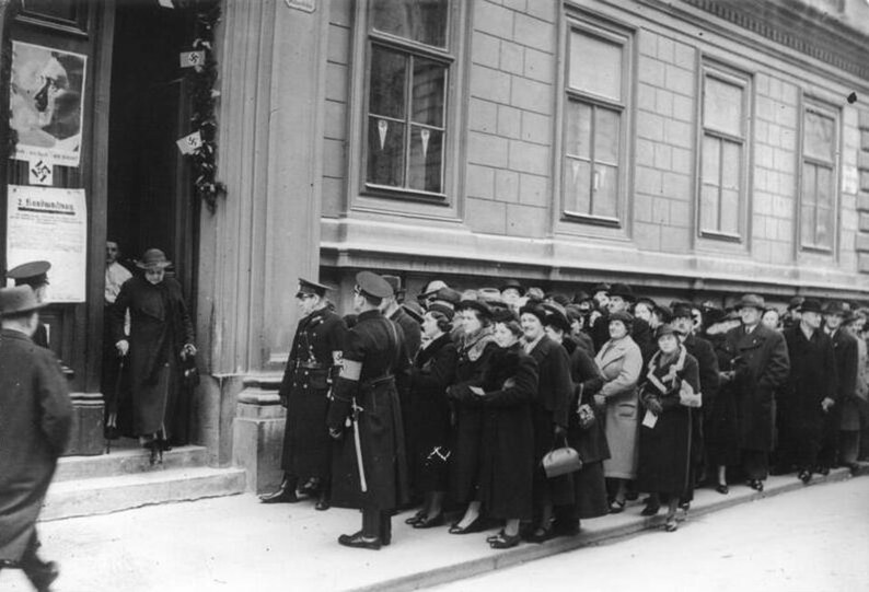 Photograph of a long queue of men and women in overcoats standing along a Vienna building at a polling station door, uniformed men with armbands beside them, swastika flags on the doorway and a notice reading Volksabstimmung