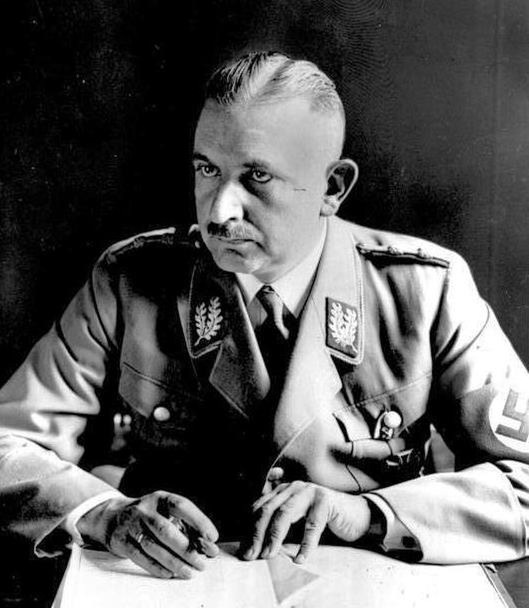 Portrait photograph of Bernhard Rust seated at a desk in a party uniform with a swastika armband, pen in hand over documents