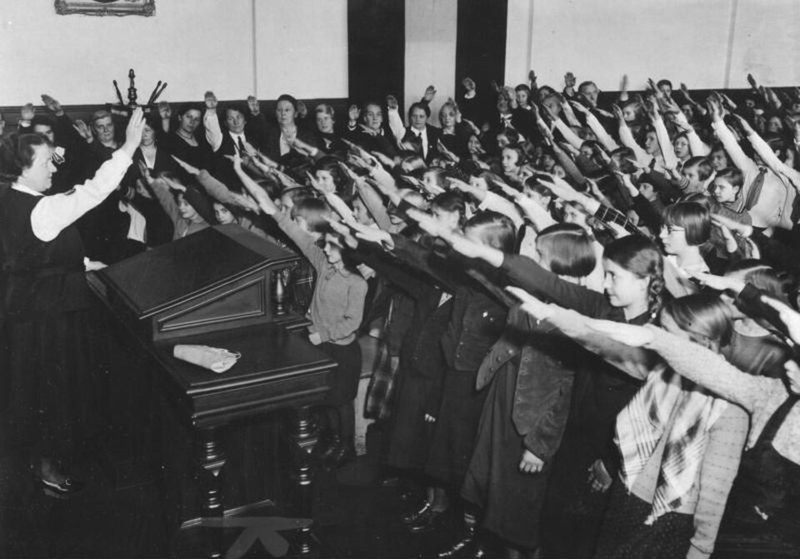 Photograph of a school class of girls and a teacher at left, all with right arms raised in the Hitler salute, in a classroom with a teacher's desk