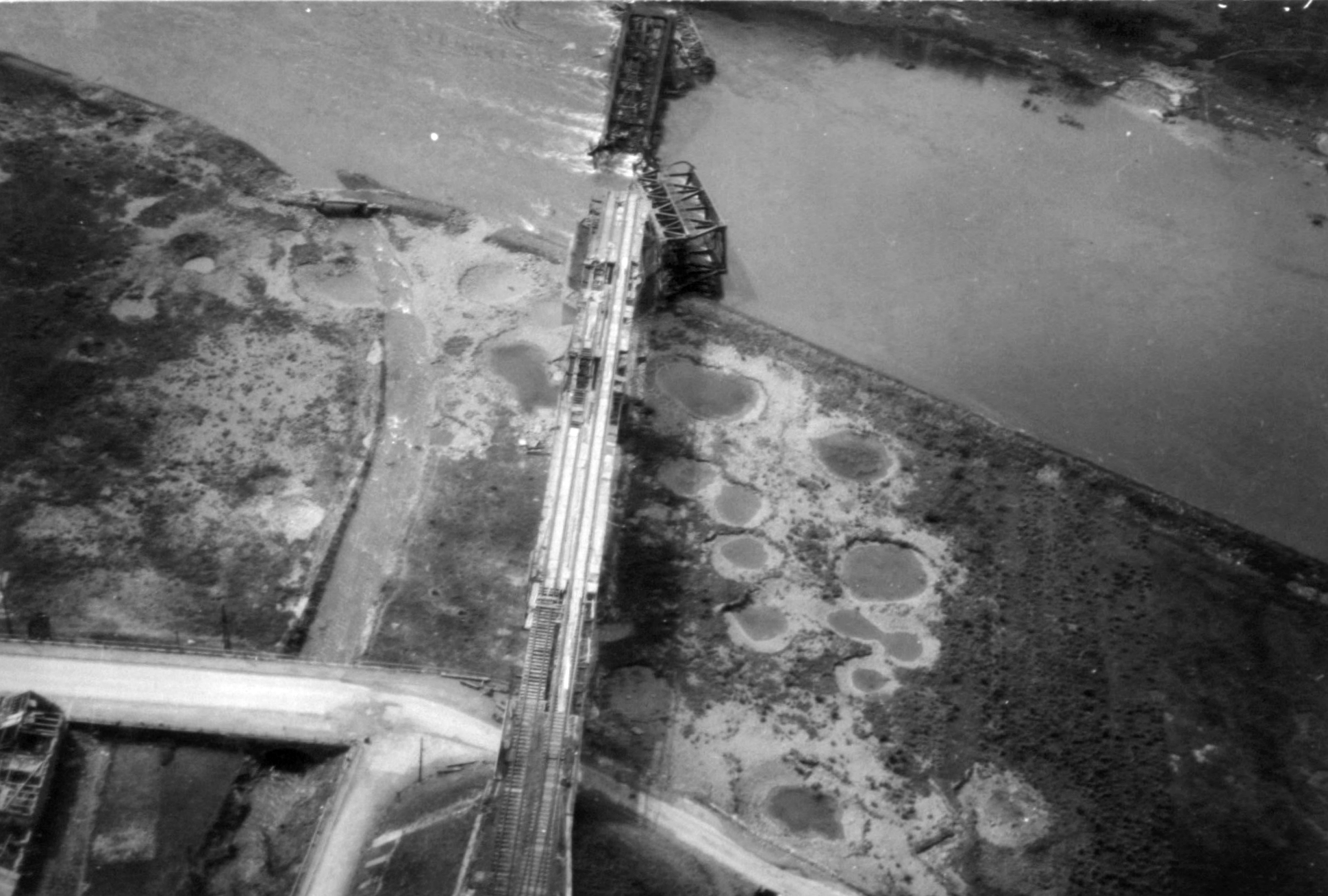Aerial view of a railway bridge at Eller with a broken span in the river, bomb craters in the fields beside it