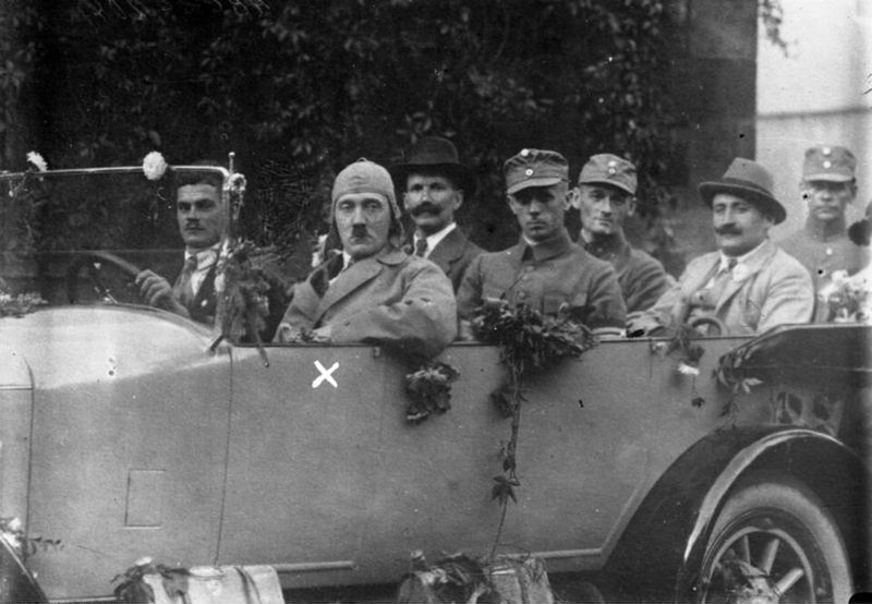 Photograph of Adolf Hitler in a cloth cap and coat, marked with a white cross, seated in a decorated open touring car with men in uniform and civilian dress