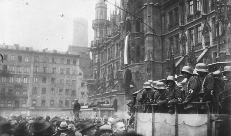 Photograph of armed men in steel helmets with swastika armbands standing on a vehicle at Munich's Marienplatz, with a dense crowd in hats in the foreground and the Rathaus tower behind
