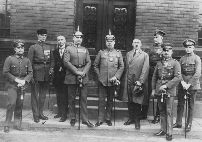 Photograph of nine men in uniform and civilian dress, including Erich Ludendorff in a spiked helmet and Adolf Hitler in a trench coat, standing in front of a courthouse door